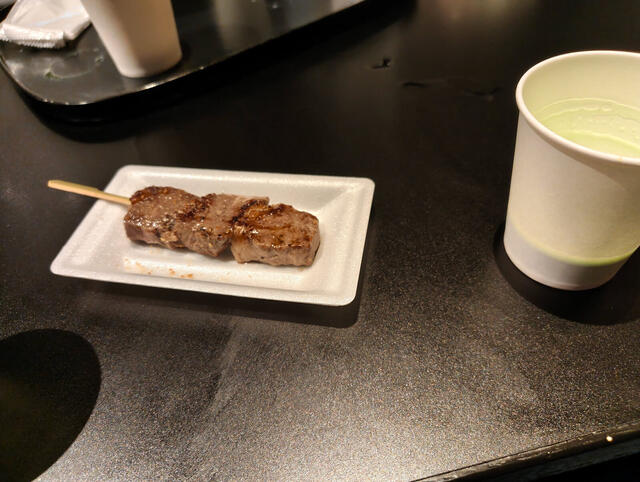 Brochette de carne grillada en bandeja de espuma y un vaso de te verde frio, de pie junto a la barra. Cerca del mercado Nishiki.